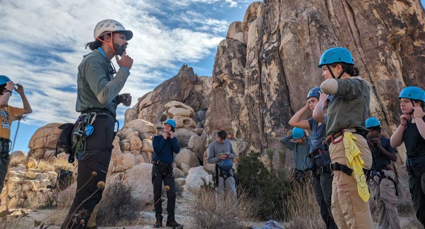 A group of students mimc the move of an instructor, who is teaching them how to properly secure a climbing helmet. They are all standing at the bottom of a tall rock wall. 